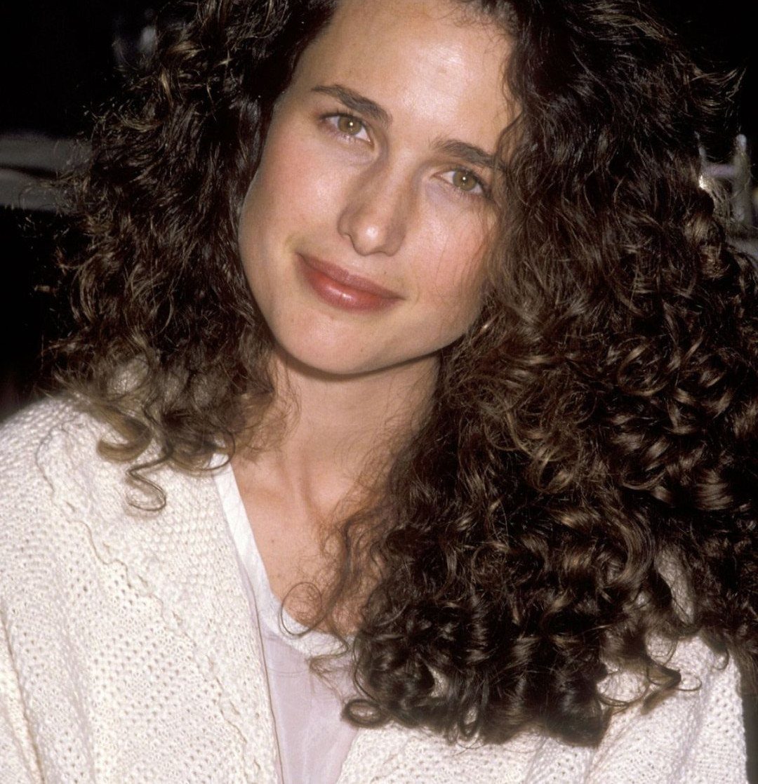 Woman with voluminous dark brown curly hair wearing a white knitted cardigan.