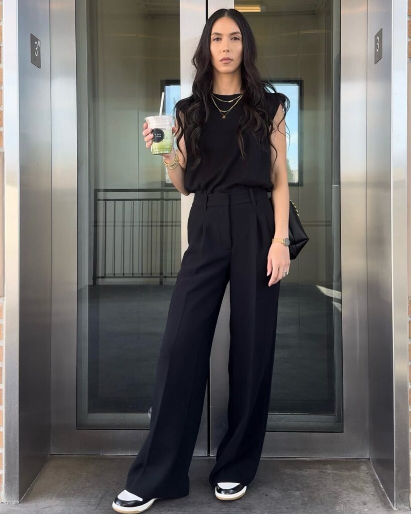 Woman wearing black wide-leg trousers and a black sleeveless top holding a drink.