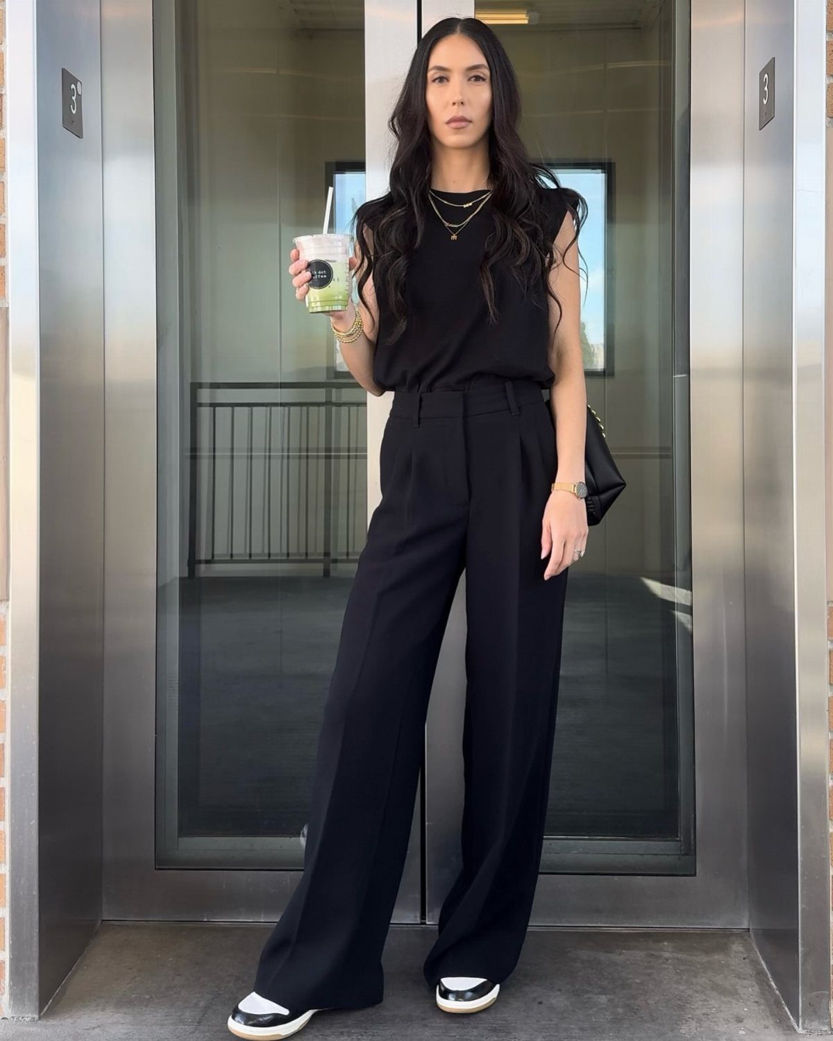Woman wearing black wide-leg trousers and a black sleeveless top holding a drink.