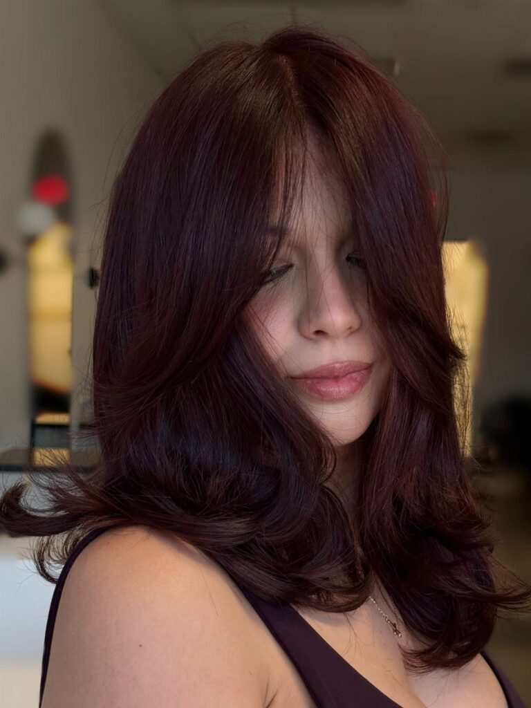 Woman with long, wavy auburn burgundy hair posing in a soft indoor setting.