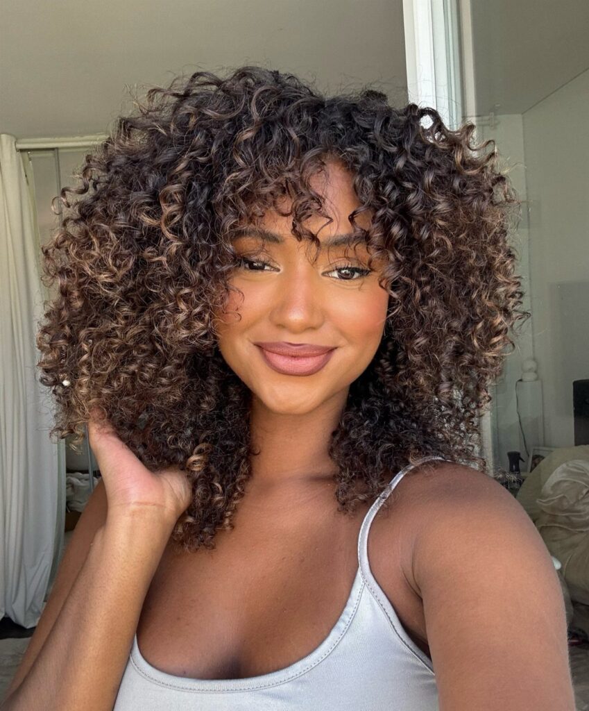 Woman with voluminous curly brown hair and golden highlights smiling in a room