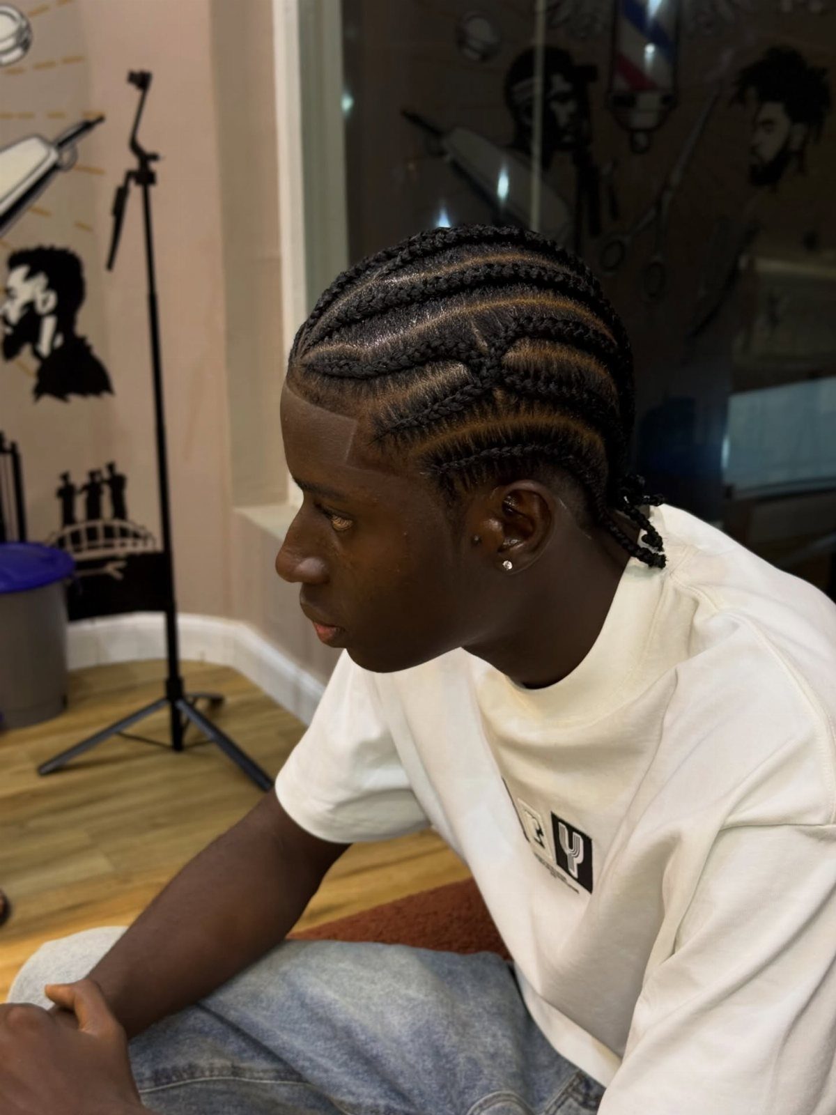 Man with cornrow braids in a barbershop wearing a white t-shirt and jeans