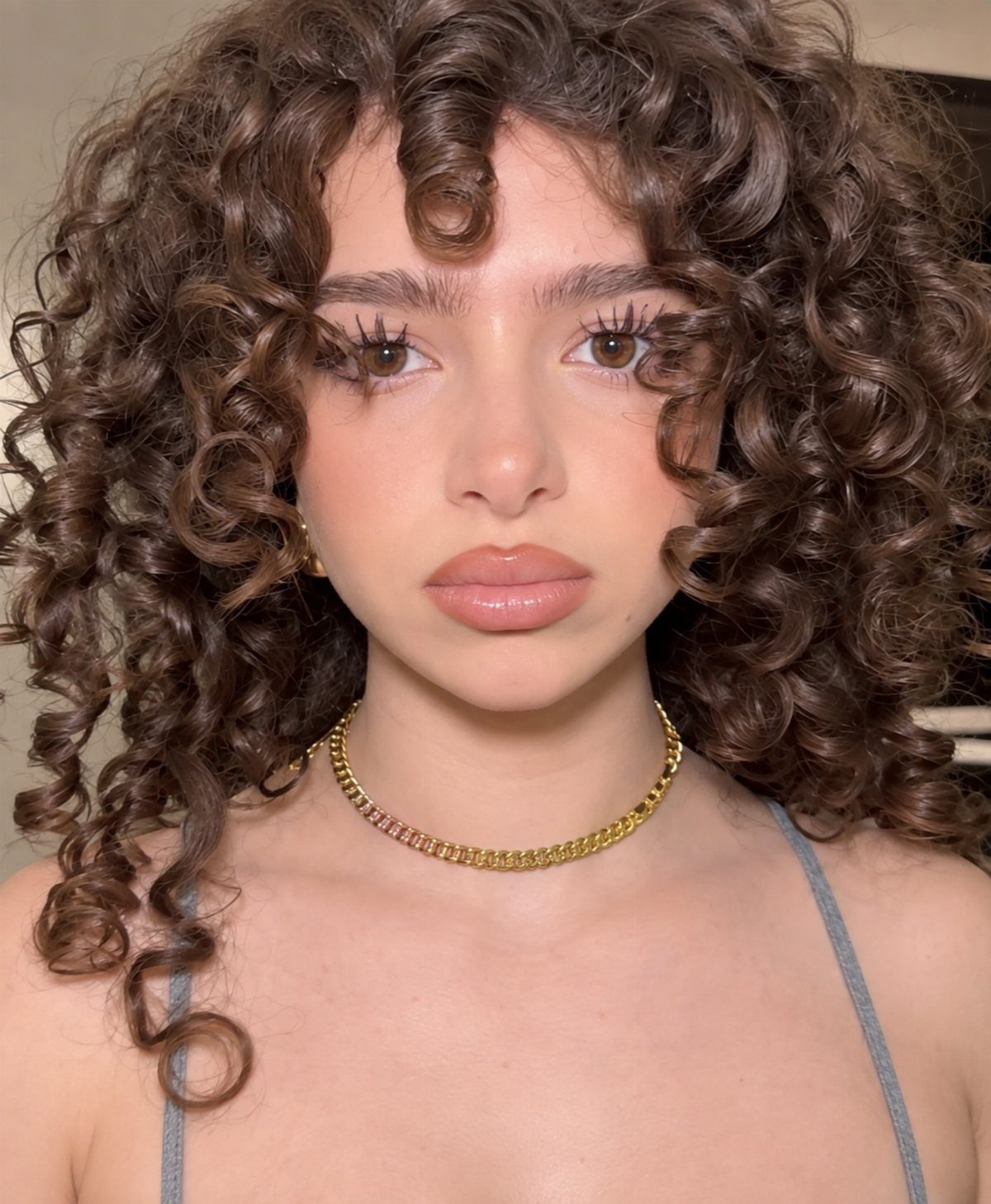 Young woman with voluminous brown curly hair and a gold chain necklace.