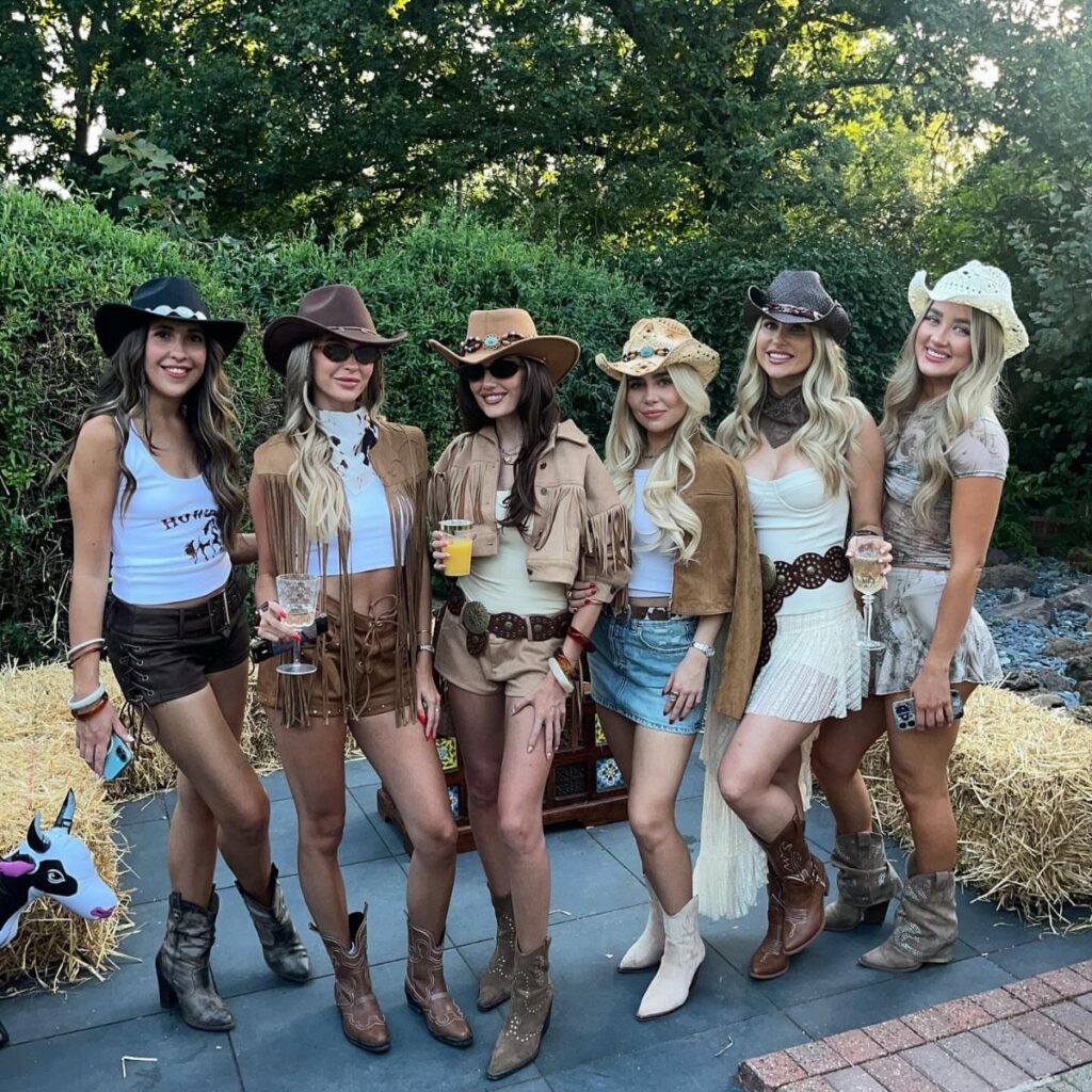 Six women wearing cute cowboy outfits with hats and boots posing outdoors.