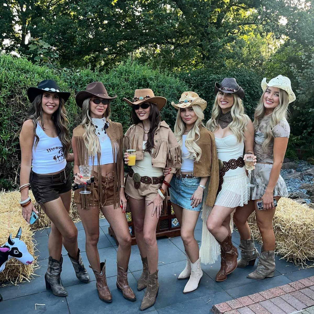 Six women wearing cute cowboy outfits with hats and boots posing outdoors.