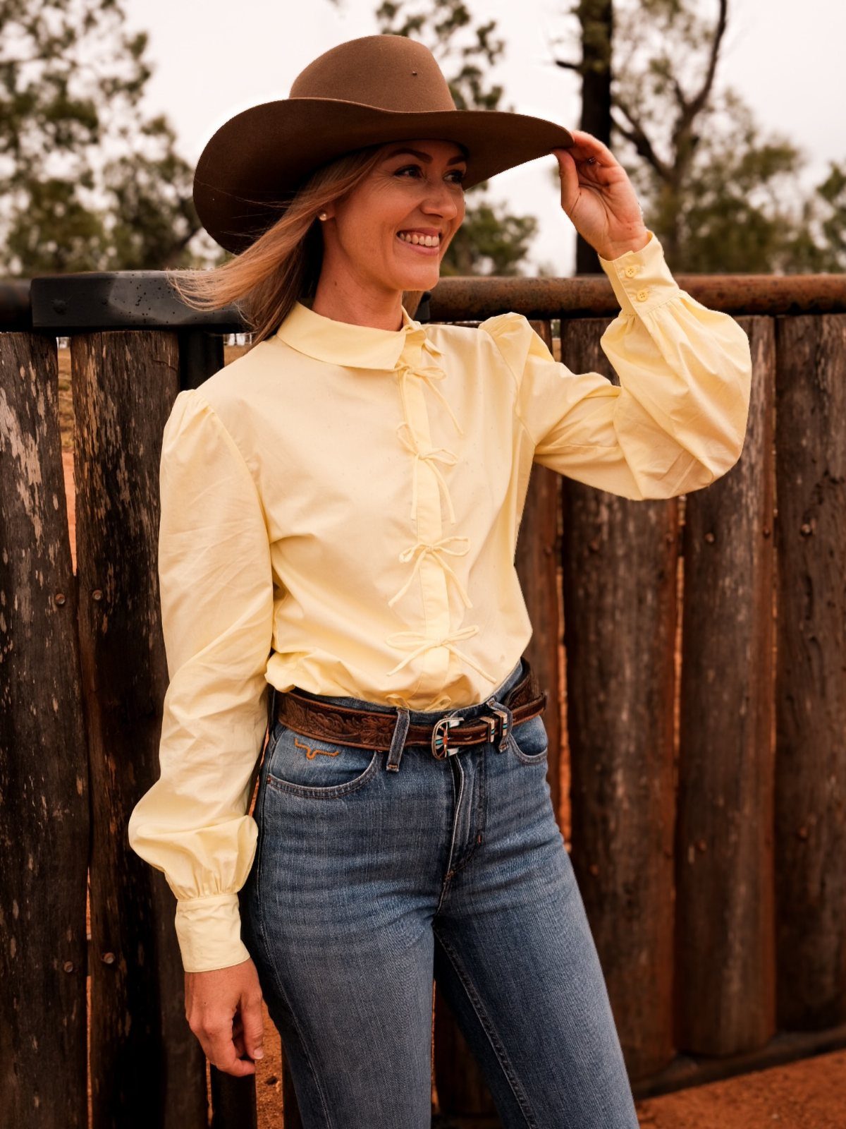 Woman in yellow western shirt, brown cowboy hat, and blue jeans by wooden fence