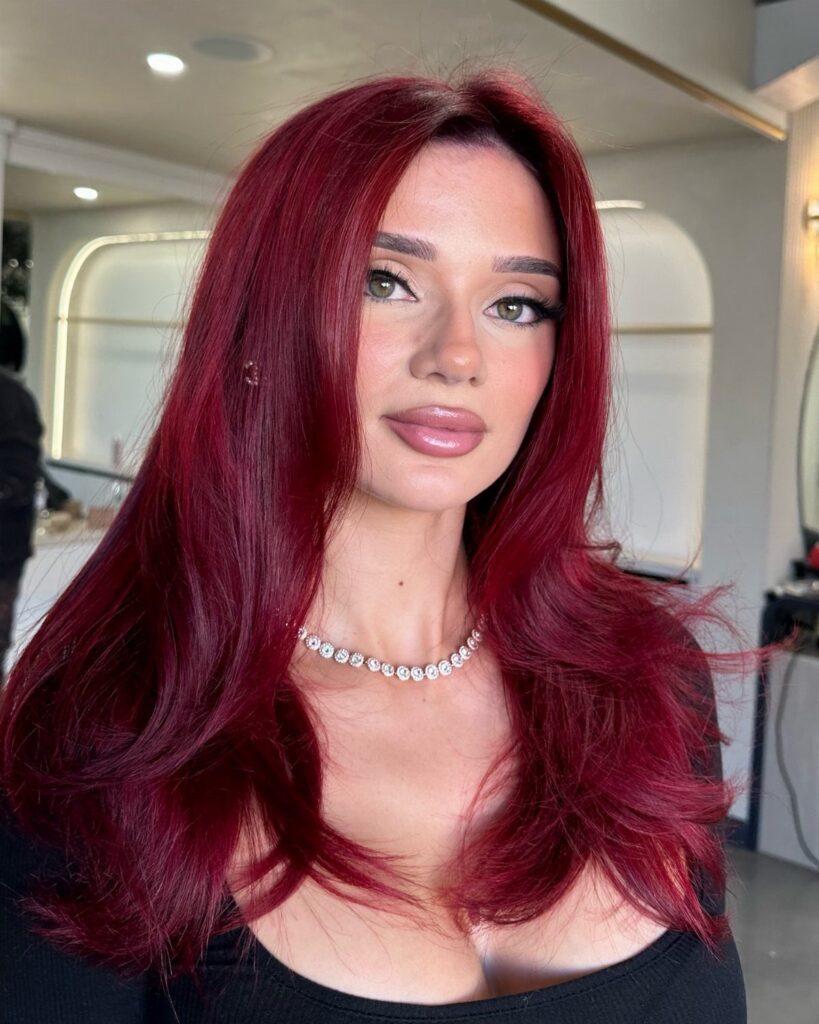 Woman with long wavy dark crimson red hair wearing a diamond necklace.
