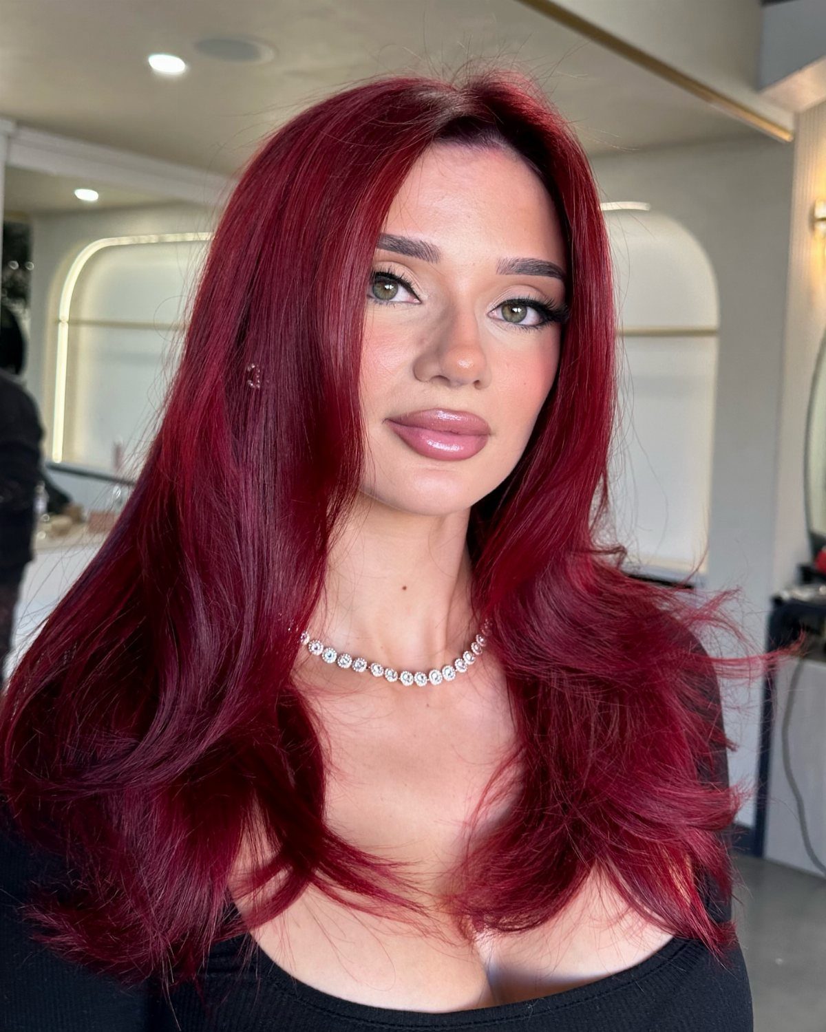 Woman with long wavy dark crimson red hair wearing a diamond necklace.