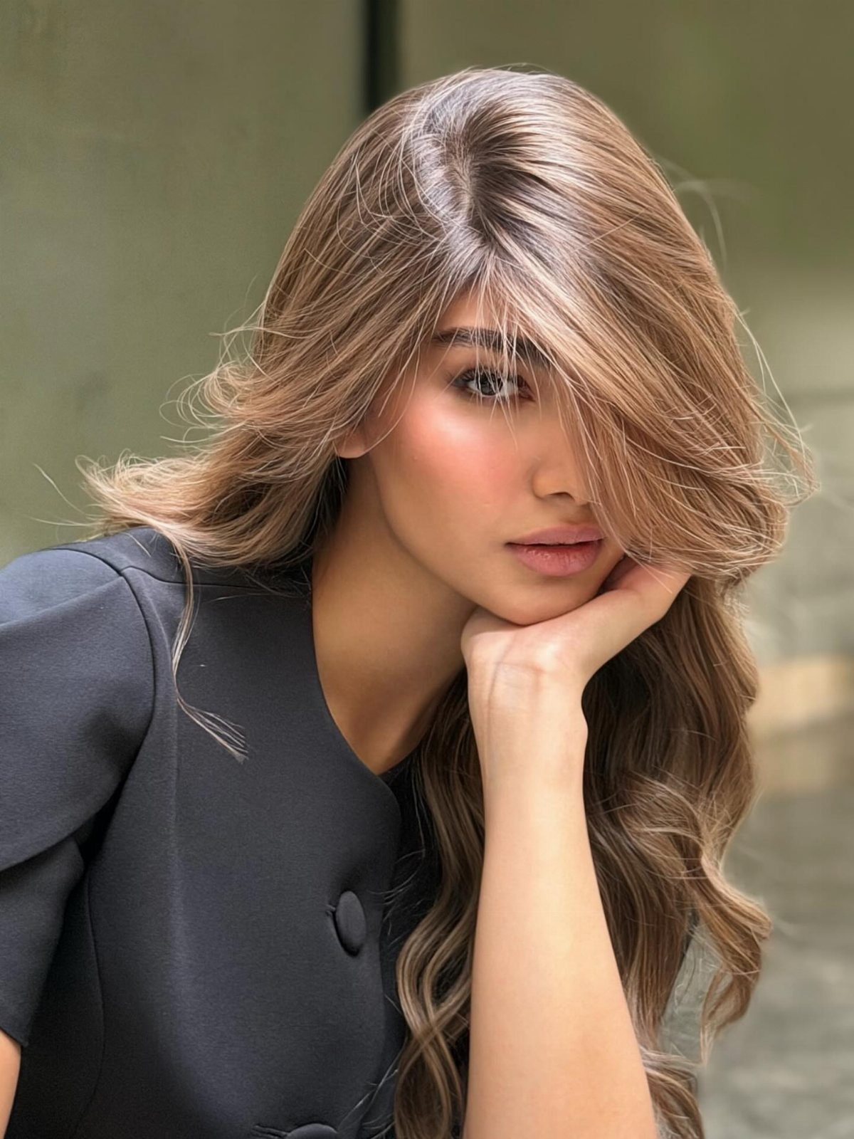 Woman with long wavy brunette hair and golden highlights wearing a black blazer.