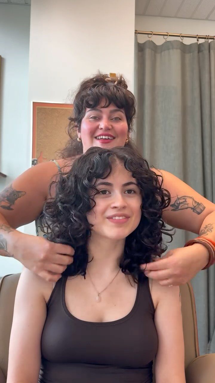 Woman with dark curly shoulder-length hair smiling while a stylist touches her curls.