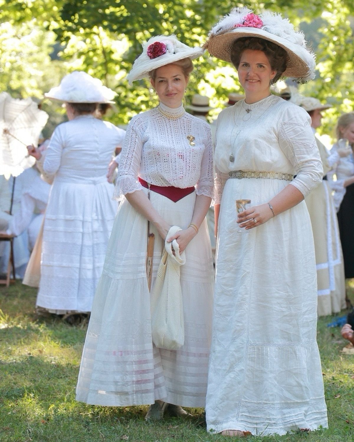 Two women wearing white Edwardian dresses and wide-brimmed hats in a grassy field.