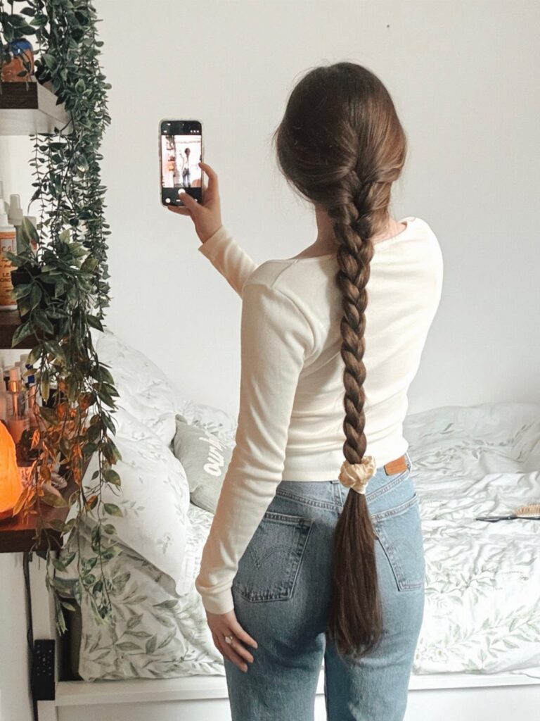 Woman with a long braided ponytail taking a mirror selfie in her bedroom.