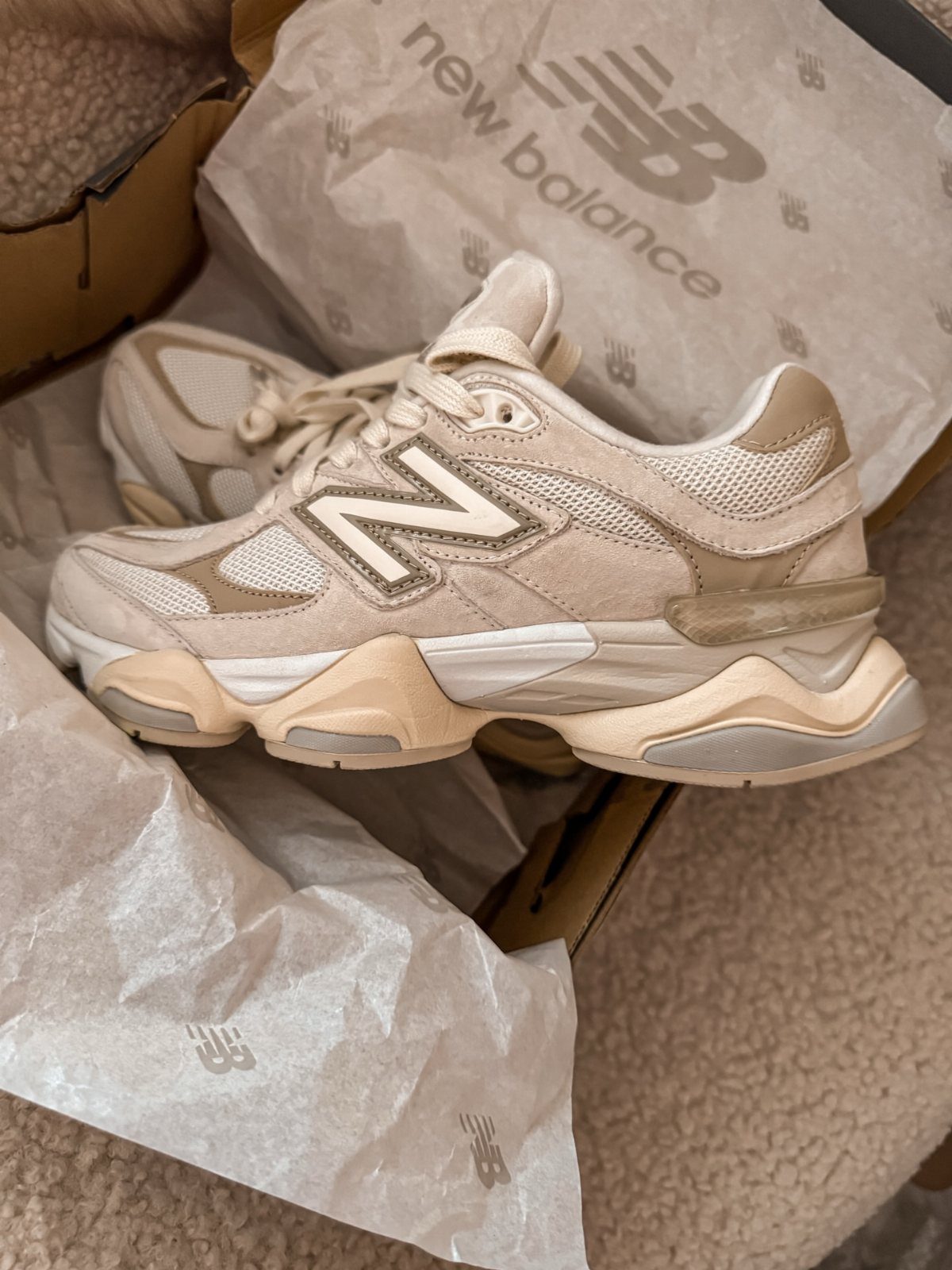 Beige New Balance sneakers sitting inside a cardboard shoebox with branded tissue paper.