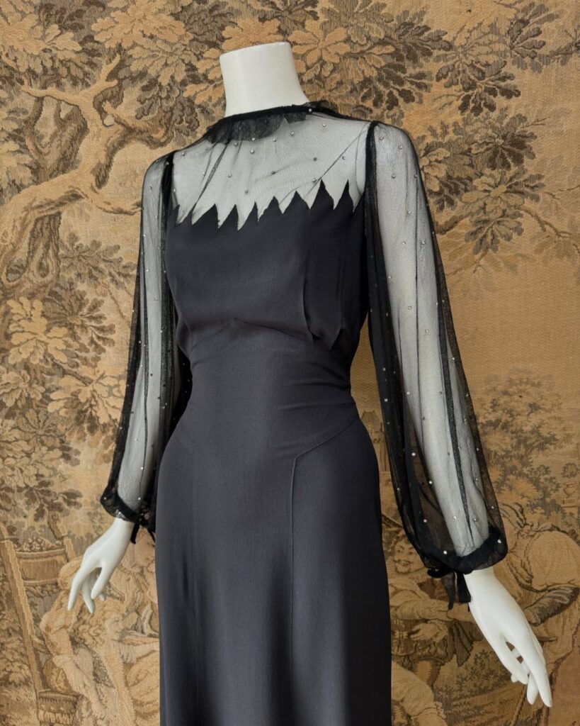 Black vintage dress with sheer beaded sleeves on a white mannequin against tapestry