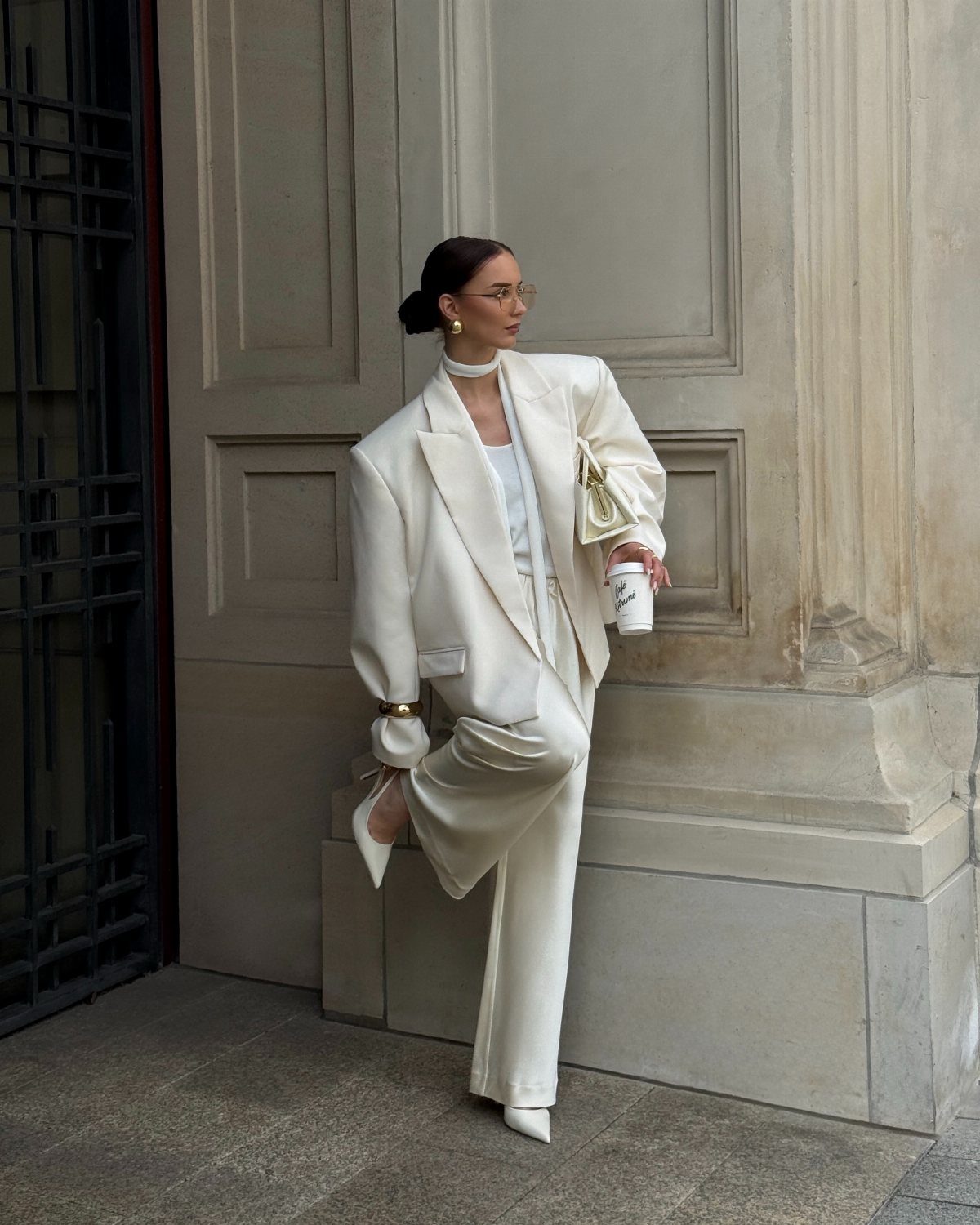 Woman in an oversized white blazer and trousers leaning against a stone wall.
