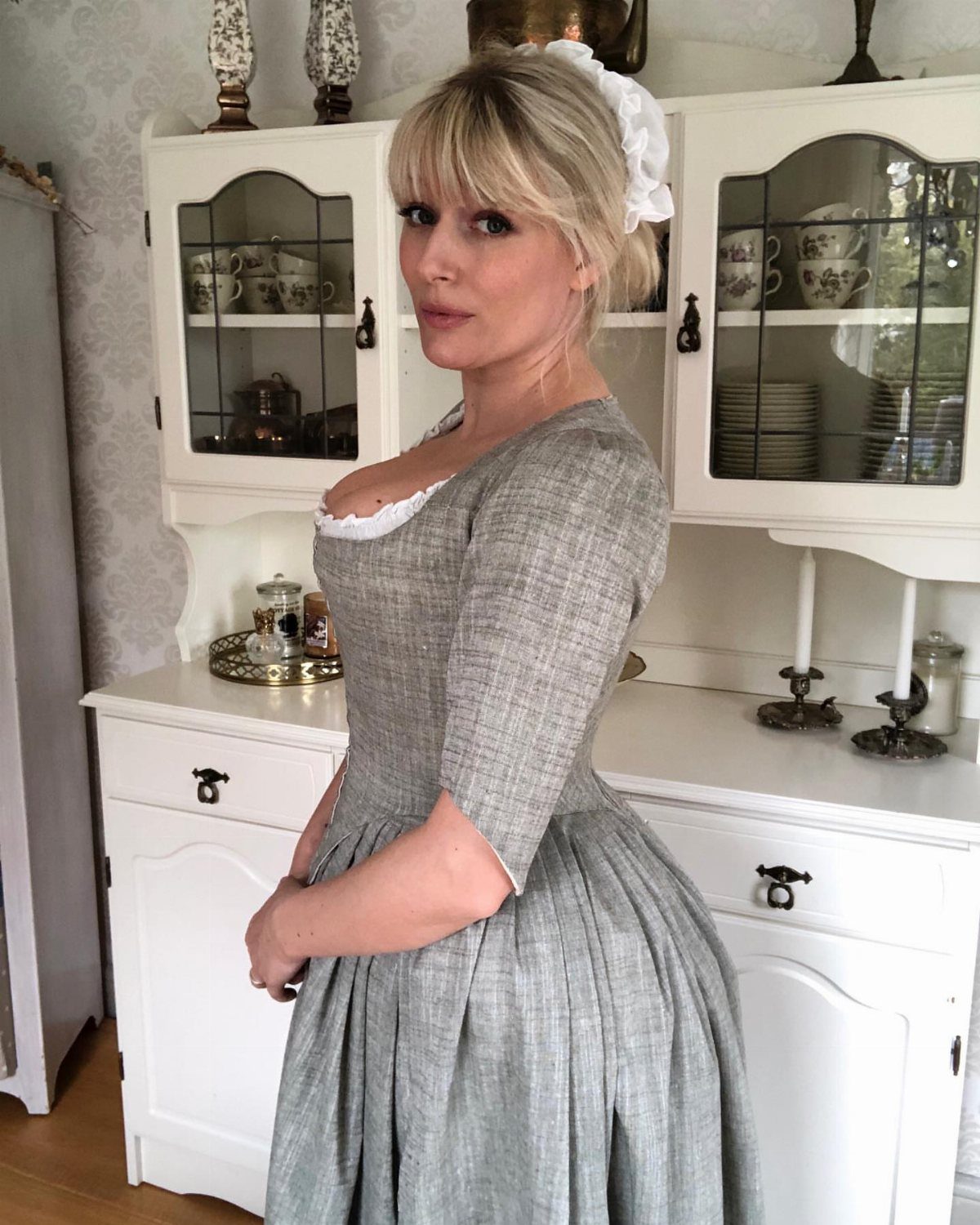 Woman in a grey linen period dress and white cap standing by a hutch