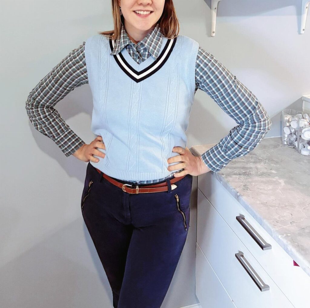 Person wearing a blue plaid shirt under a light blue sweater vest and navy trousers.