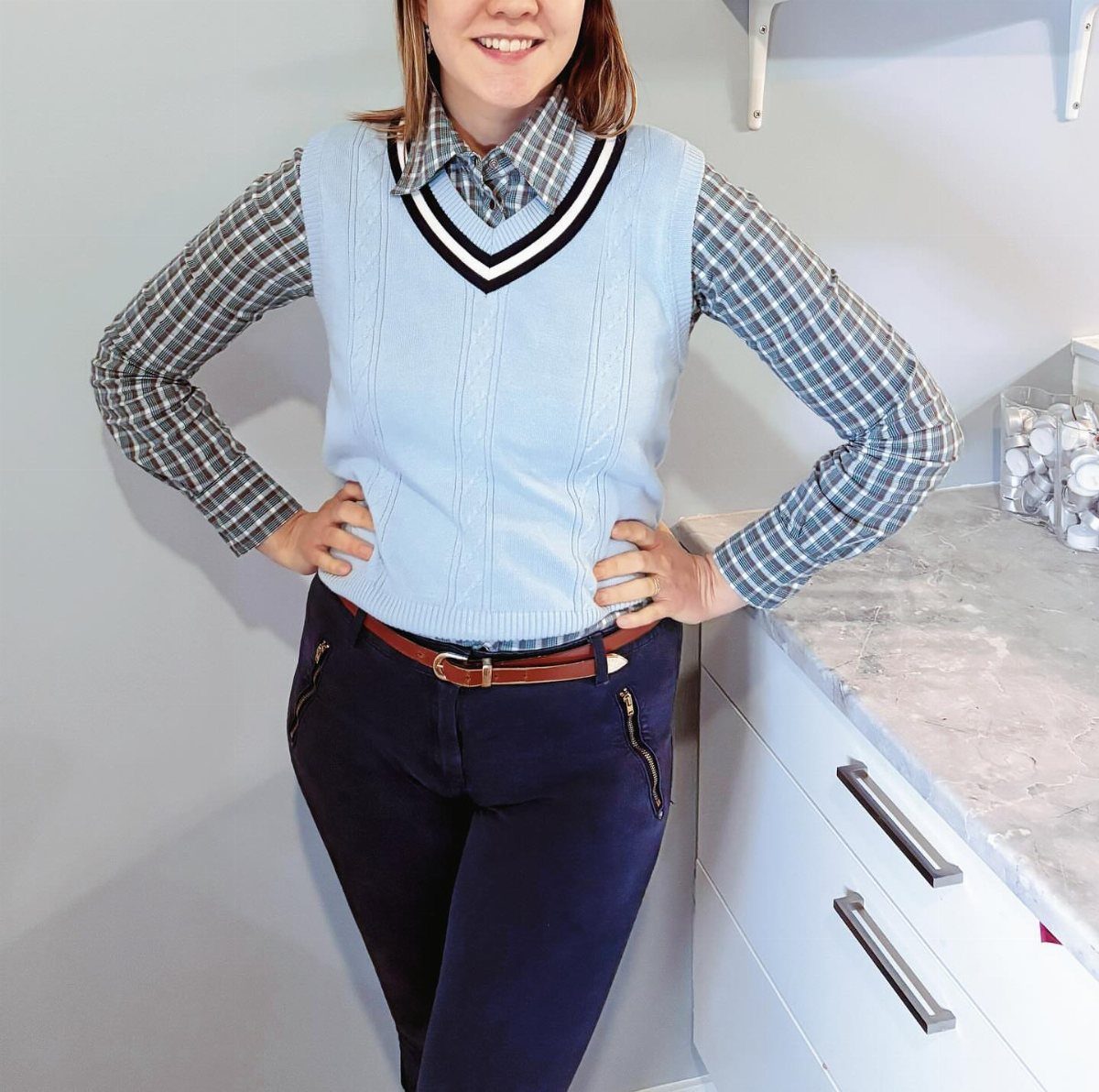 Person wearing a blue plaid shirt under a light blue sweater vest and navy trousers.